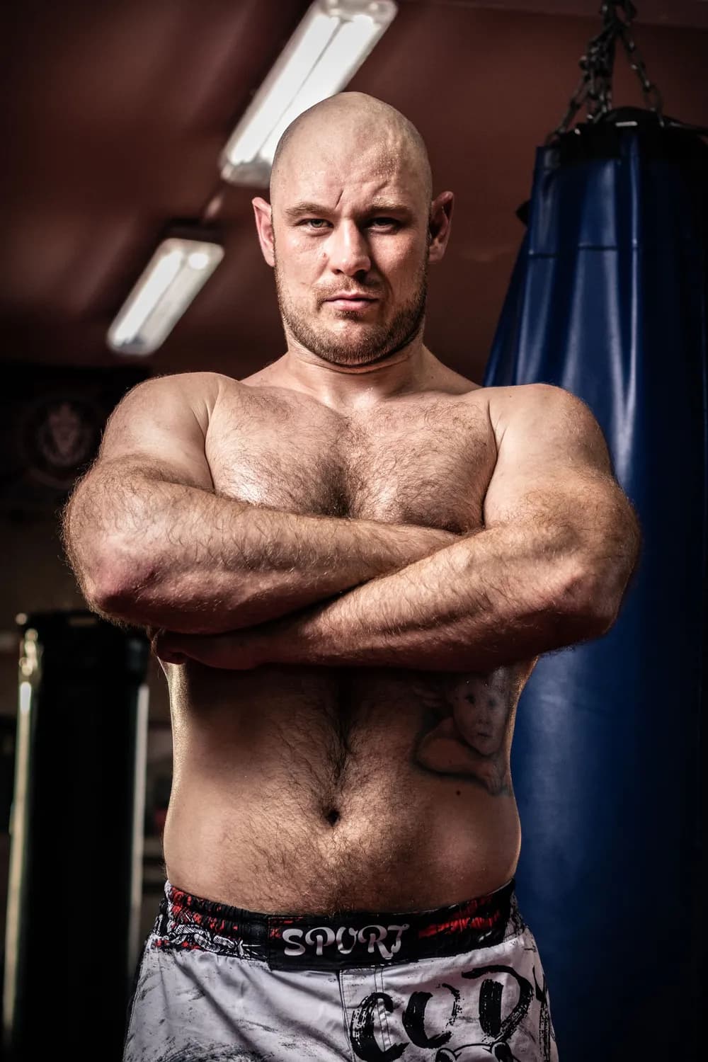 A bald, bearded coach with his arms folded in a dim boxing gym