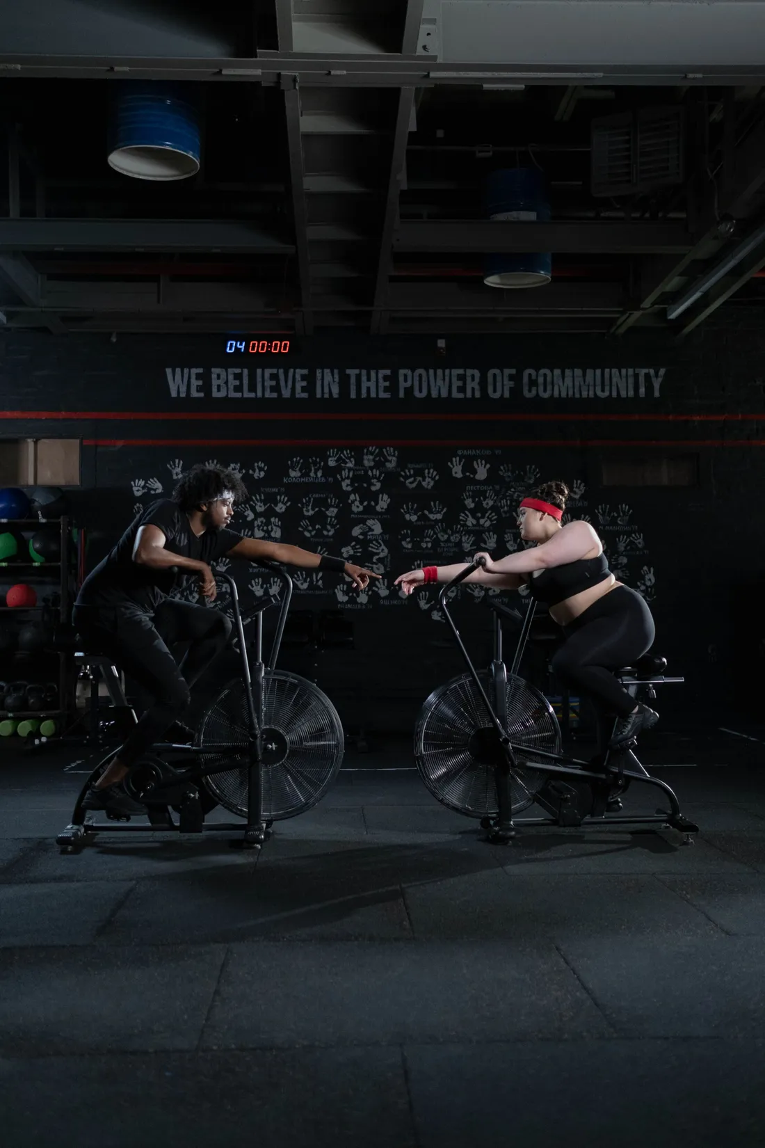 Two members on air bikes under a wall of medicine balls