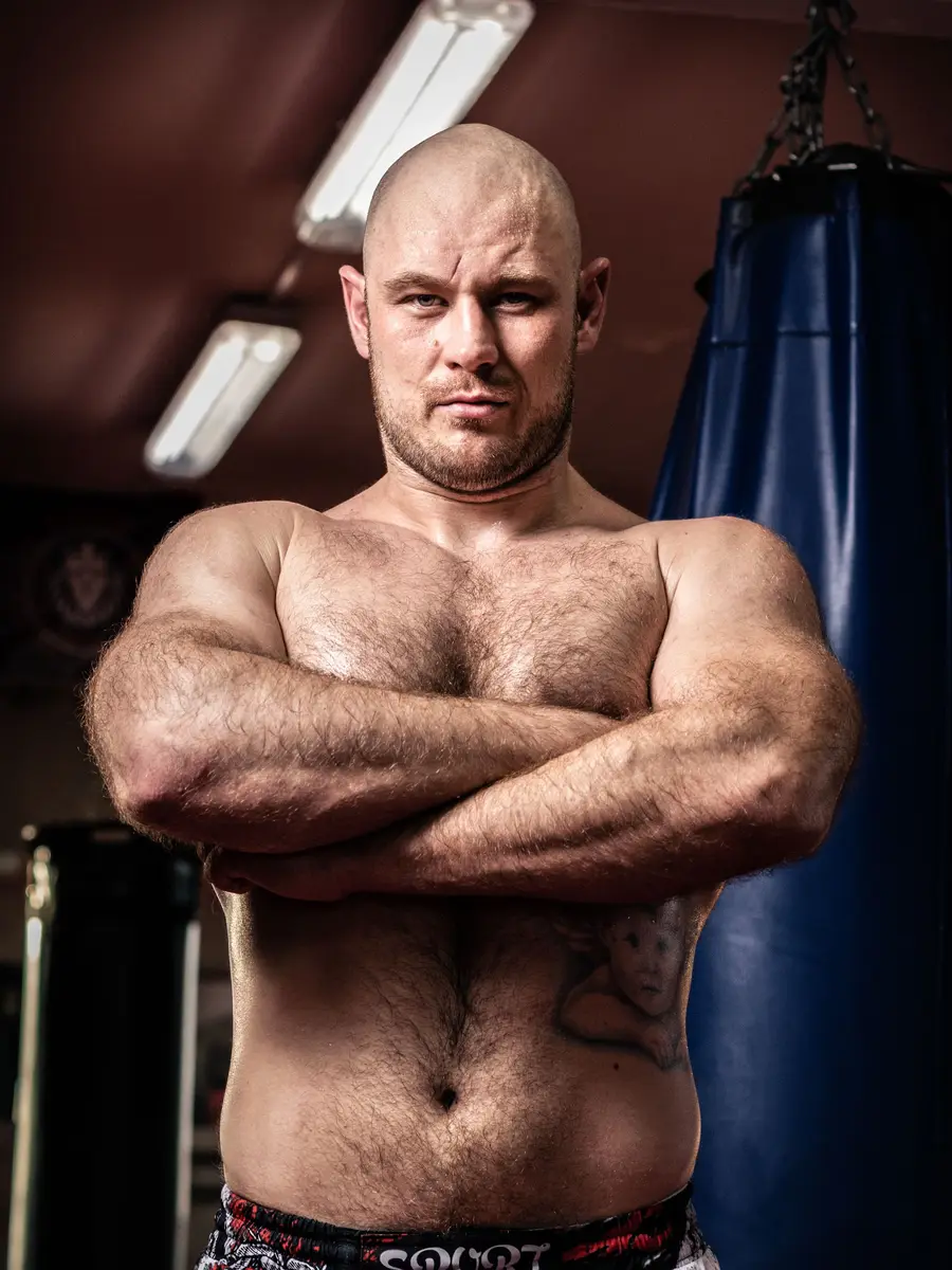 A boxing coach with folded arms beside a heavy bag under a strip light