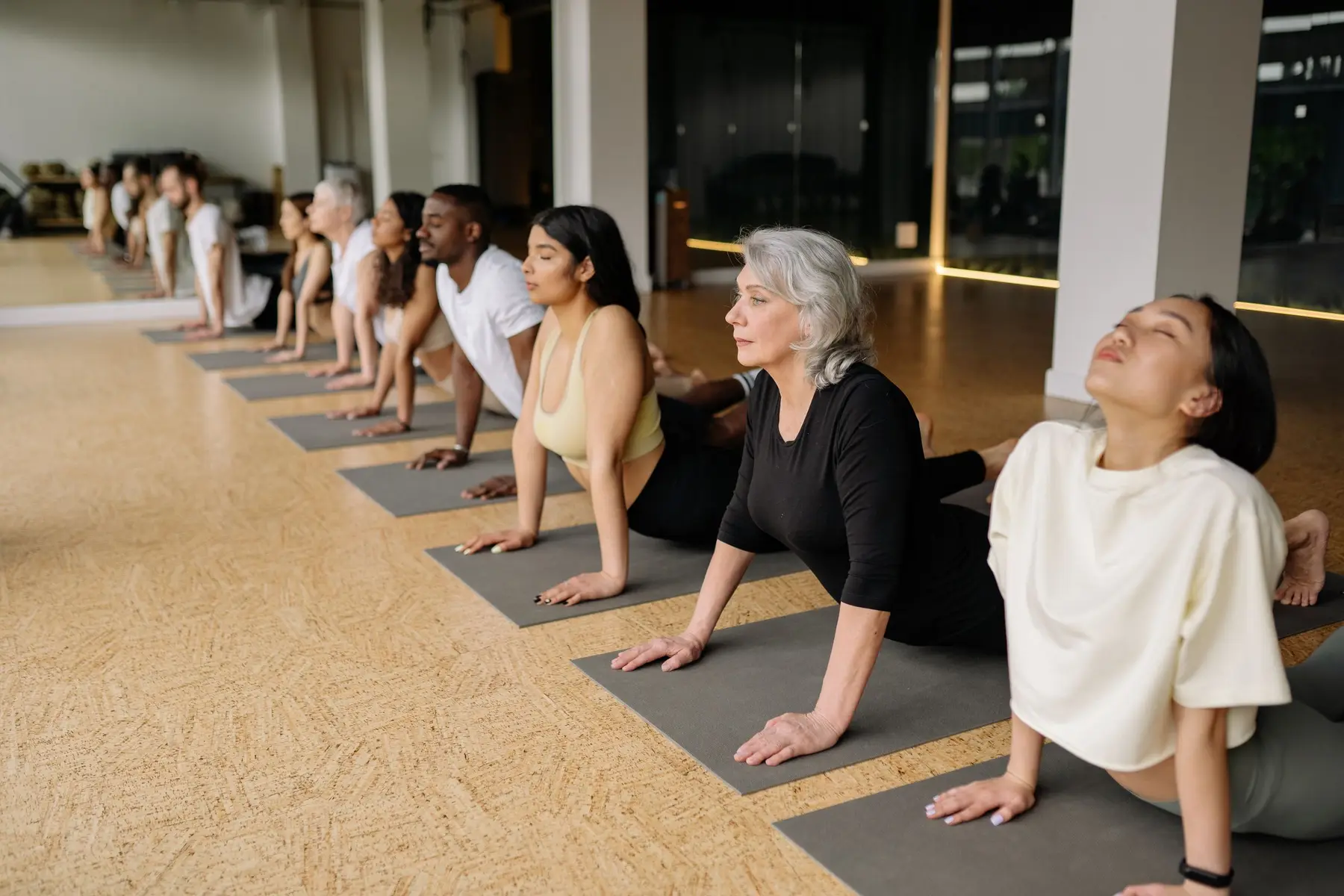 A bright studio with a long row of people stretching on mats in morning light