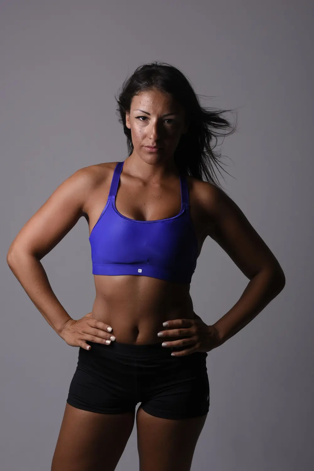An athlete in a blue sports top, hands on hips, lit from one side against a grey studio wall