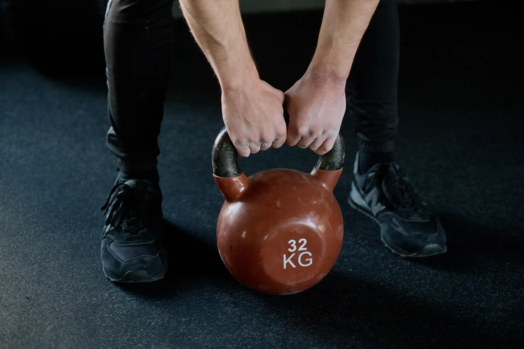 Hands and trainers either side of a red 32 kg kettlebell on a dark floor