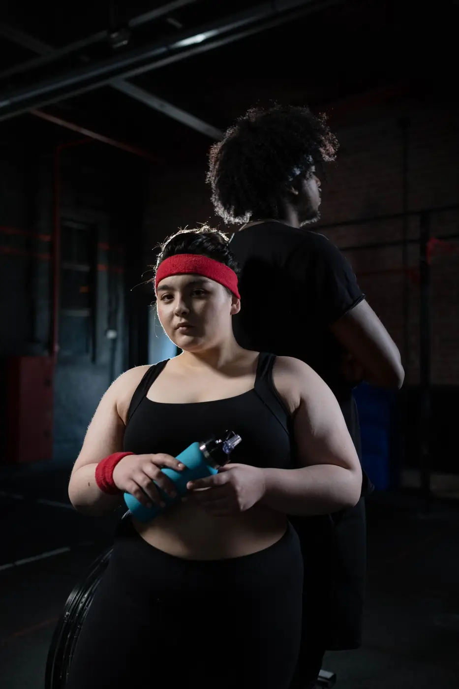 A woman in a red headband holding a water bottle in a dark gym