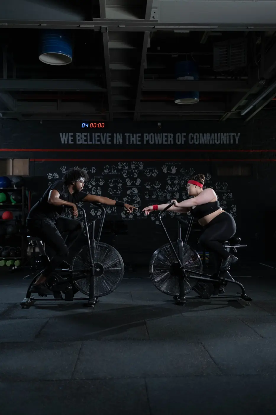 Two people on air bikes in a dark room under a slogan on the wall