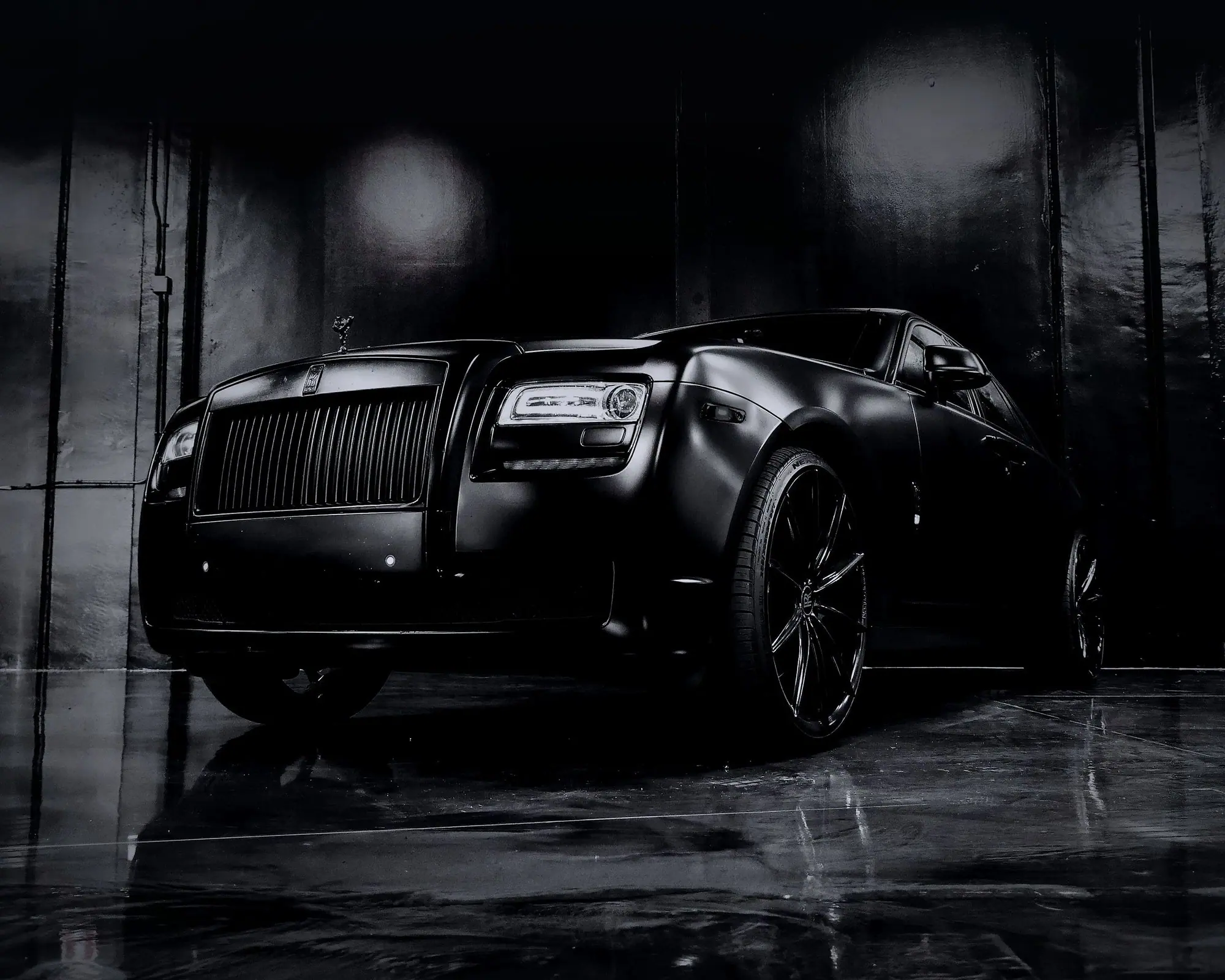 A matte black luxury saloon in a dark studio, seen low from the front three-quarter on a wet floor