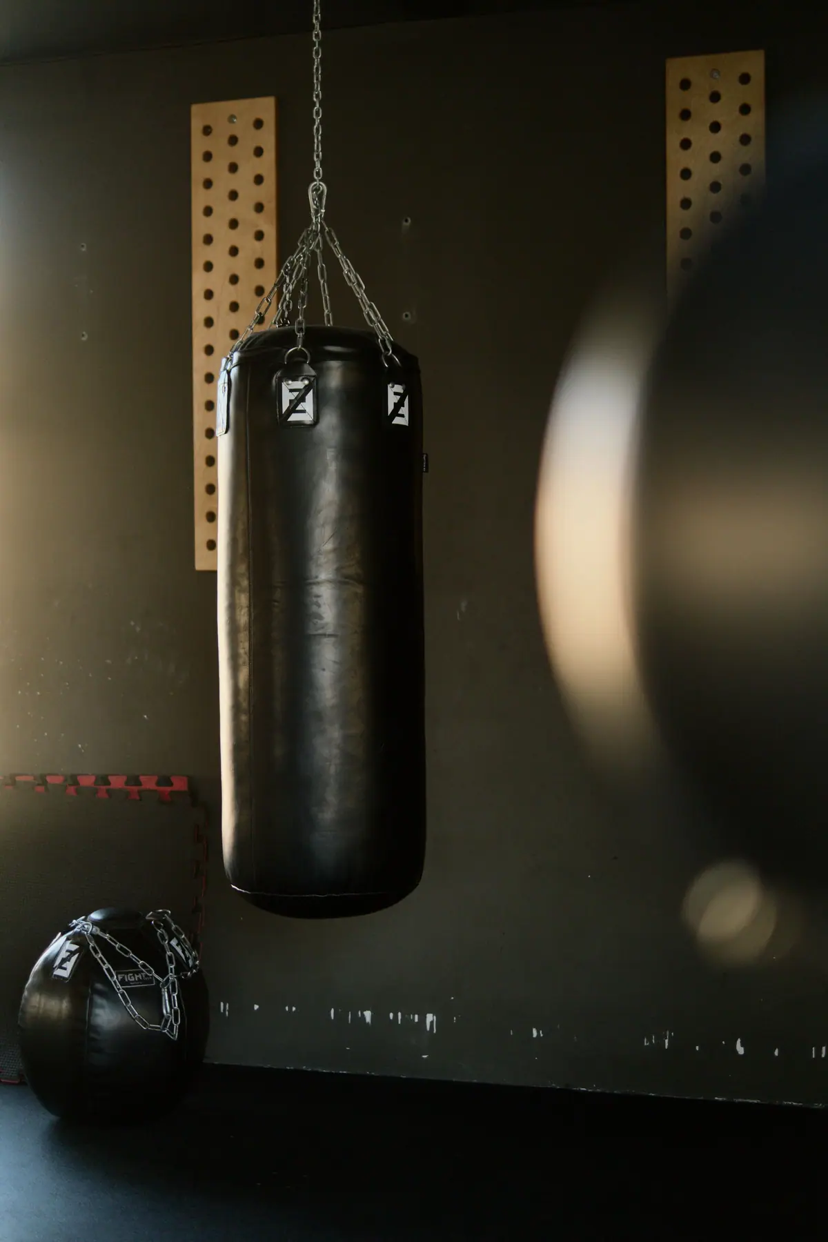 A black punching bag hanging on a chain in a dim gym, a medicine ball on the floor beneath