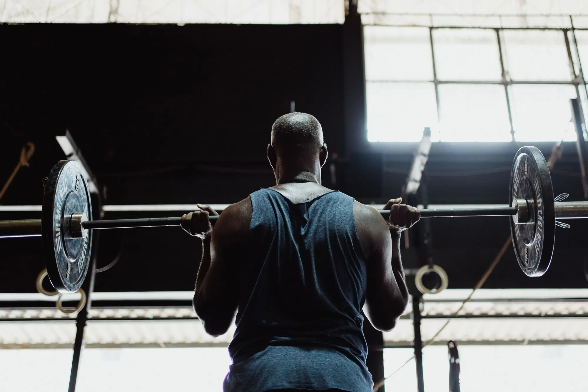 An athlete seen from behind holding a loaded barbell at chest height, backlit by a bright industrial window