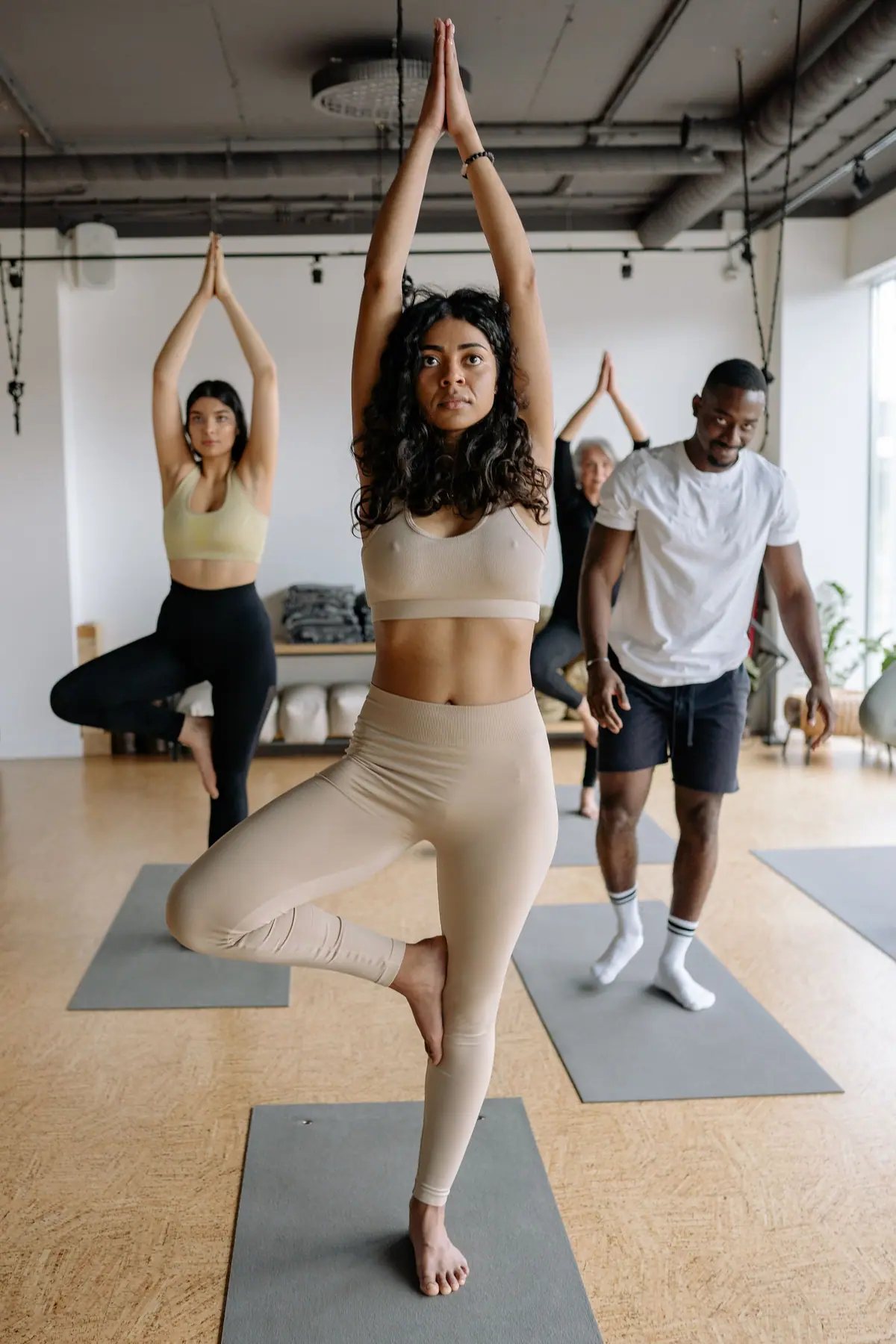 A yoga class in a bright studio, a woman in front balancing in tree pose with her hands raised